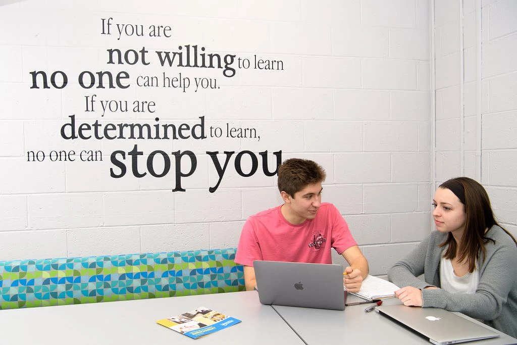 Two people are studying together at a table, with an inspirational quote about learning on the wall behind them.